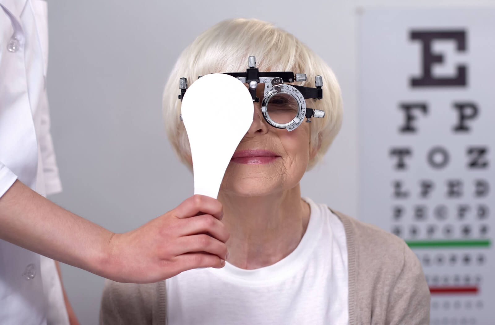 A senior woman in an eye exam wearing a trial frame device while an optometrist covers her right eye