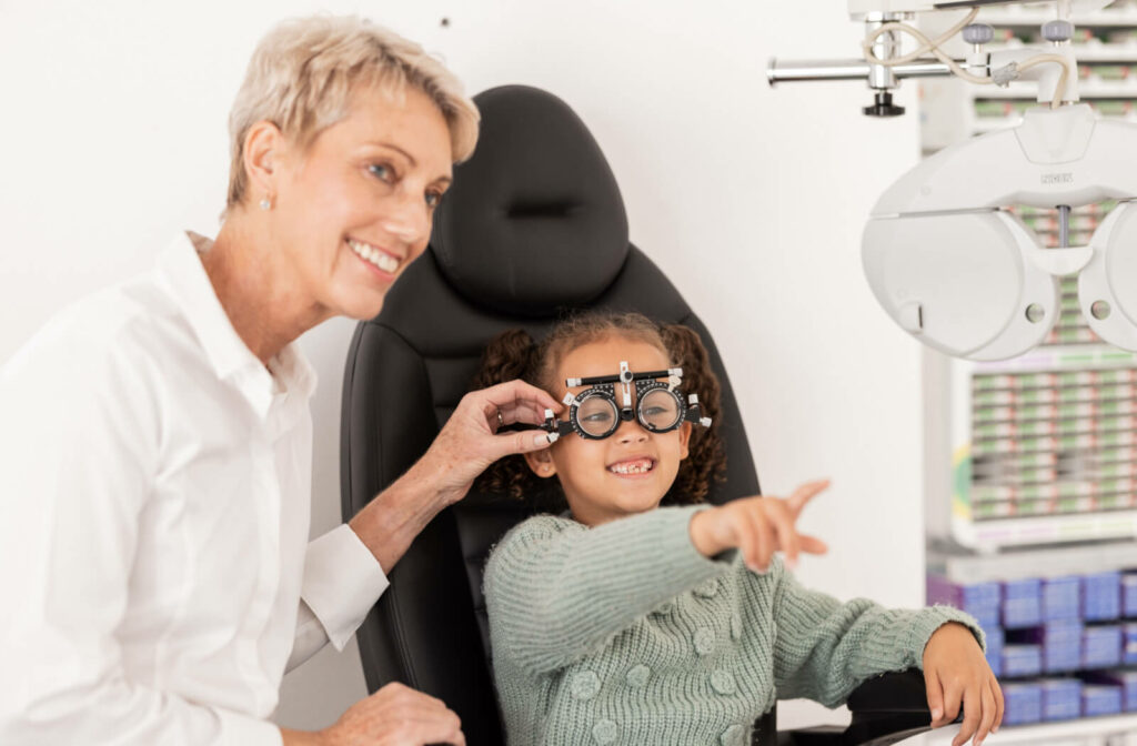 A young girl smiling and pointing ahead while a female optometrist checks her measurements for glasses