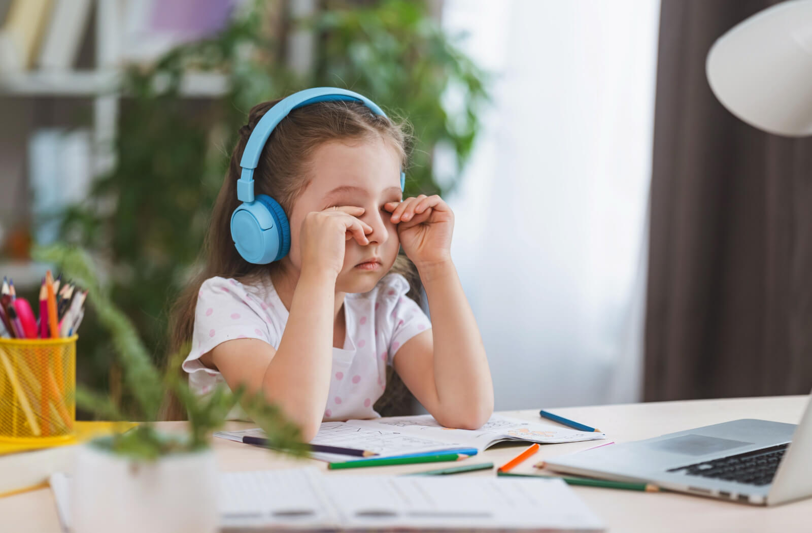 A young girl wearing blue headphones sits at a table with a book, coloured pencils, and a laptop. She is rubbing her eyes with both hands.