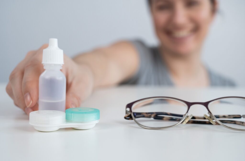 A pair of eyeglasses and a pair of contact lenses sit on a table. An adult reaches out to grab the bottle of contact lens solution sitting behind the contacts.
