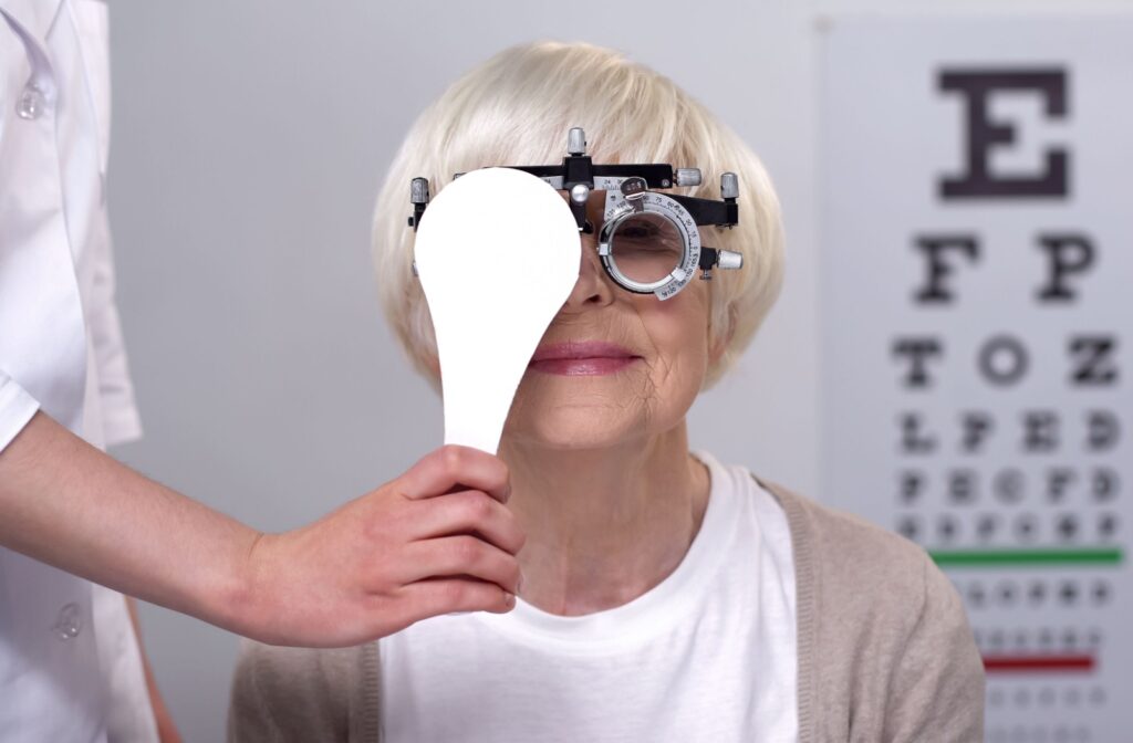A senior woman in an eye exam wearing a trial frame device while an optometrist covers her right eye.