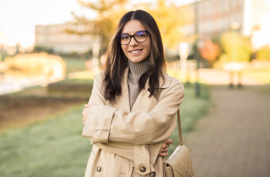 A young adult standing outside in a trench coat wearing glasses.